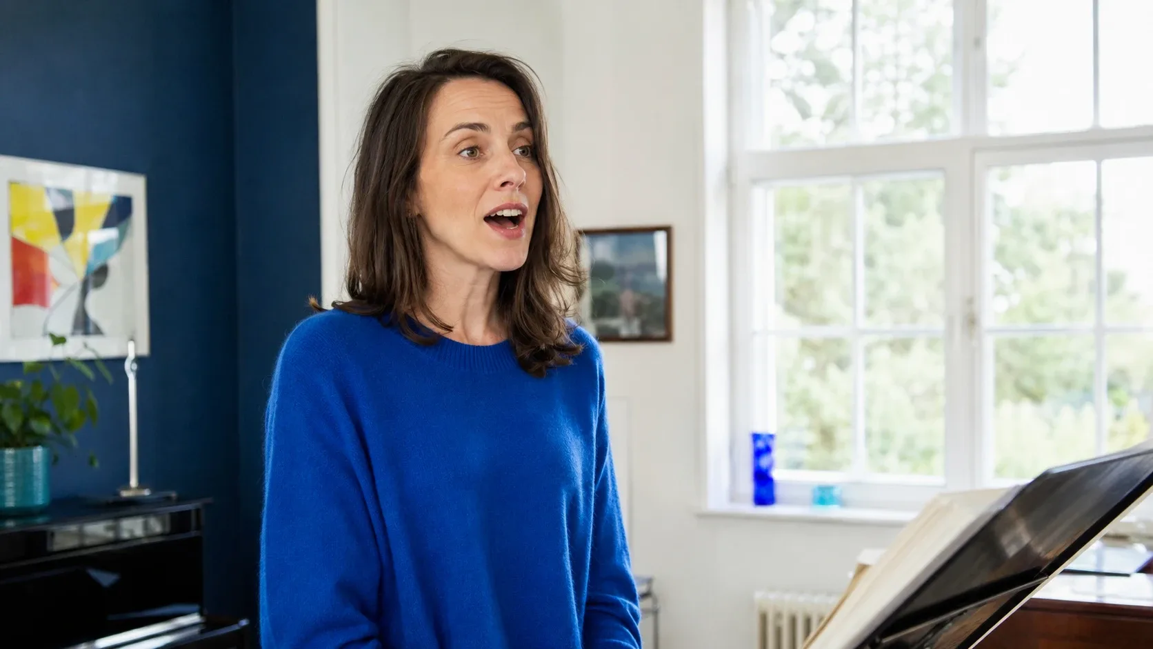 A woman in a bright blue sweater singing in a light-filled practice room, standing beside an upright piano with an open score on the music stand